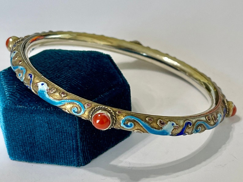K&ouml;nnte beinhalten: Ein silbernes Armreif-Armband mit blauen und wei&szlig;en Emaildetails und roten Korallenakzenten. Das Armband ist mit einem wirbelnden Muster und V&ouml;geln verziert.