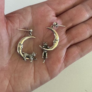 May include: A pair of silver earrings featuring a crescent moon design with a cat perched on the edge. The cat is facing the moon and has a detailed, realistic design.