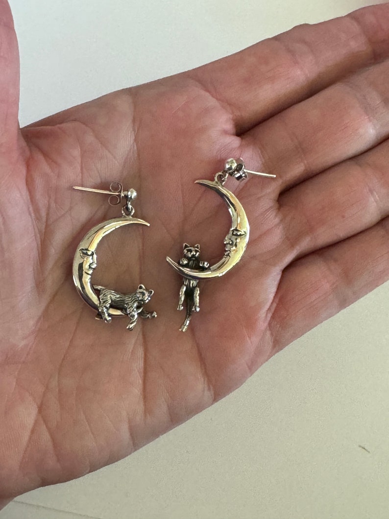 May include: A pair of silver earrings featuring a crescent moon design with a cat perched on the edge. The cat is facing the moon and has a detailed, realistic design.