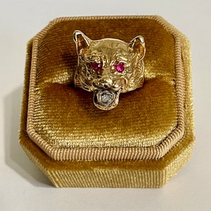 May include: A gold ring with a detailed animal head design. The animal has red ruby eyes and a diamond in its mouth. The ring is displayed on a gold velvet ring box.