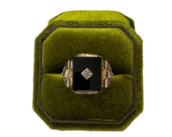 May include: A vintage ring featuring a rectangular black onyx stone and a small diamond. The ring is set in gold-toned metal and presented in a green velvet octagonal ring box. The ring is a classic design.