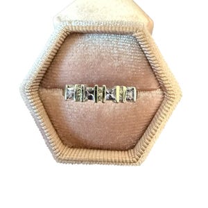 Puede incluir: Un anillo dorado con piedras rectangulares y cuadradas se muestra dentro de un joyero de terciopelo rosa. El anillo presenta un diseño lineal con piedras y segmentos metálicos alternos, creando un aspecto moderno y elegante.
