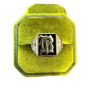 Puede incluir: Un anillo de plata con un centro de ónix negro que presenta la letra "R" estilizada en una fuente decorativa. El anillo se exhibe en un joyero de terciopelo verde brillante.