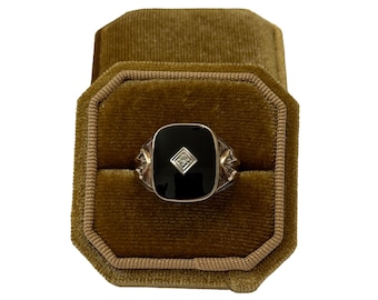 May include: A silver ring featuring a black onyx square centre stone and a small diamond. The ring is presented in a brown velvet ring box. The ring has decorative details on the sides.