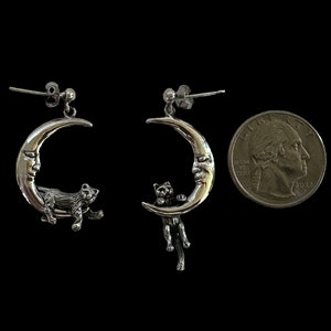 May include: A pair of silver earrings featuring a crescent moon with a face and a cat hanging from it. The earrings are about the size of a US quarter.
