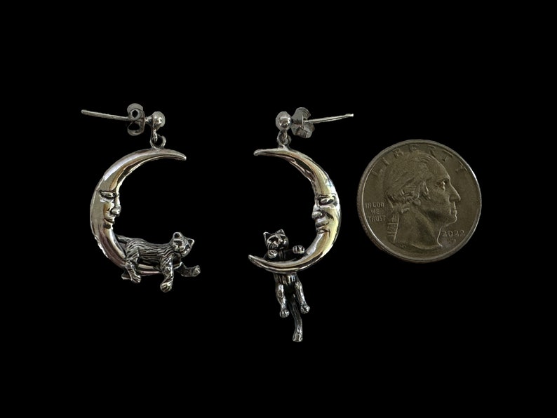 May include: A pair of silver earrings featuring a crescent moon with a face and a cat hanging from it. The earrings are about the size of a US quarter.