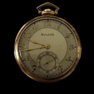 May include: A gold-tone Bulova pocket watch with a round case and a winding crown at the top. The off-white dial features gold-colored Arabic numerals, gold hands, and a smaller sub-dial at the bottom for seconds with black markings.