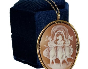 May include: A gold chain necklace with a cameo pendant featuring a carved scene of three women dancing. The cameo is set in a gold oval frame.