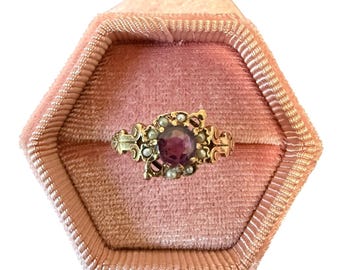 Op de afbeelding: Een goudkleurige ring met een grote, ronde, paarse edelsteen in het midden, omringd door kleinere parels en rode accenten. De ring ligt in een roze fluwelen zeshoekige ringdoos, wat een vintage uitstraling creëert.