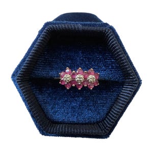 Peut inclure: Une bague en or ornée de trois motifs en forme de fleur disposés en ligne. Chaque motif se compose de six petits rubis ronds rose foncé entourant un diamant central rond et transparent. La bague est présentée dans un écrin en velours bleu.