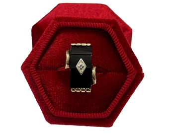 May include: A black and gold ring with a geometric design, featuring a small diamond. The ring is displayed inside a red velvet ring box. The ring has a rectangular shape with gold accents.