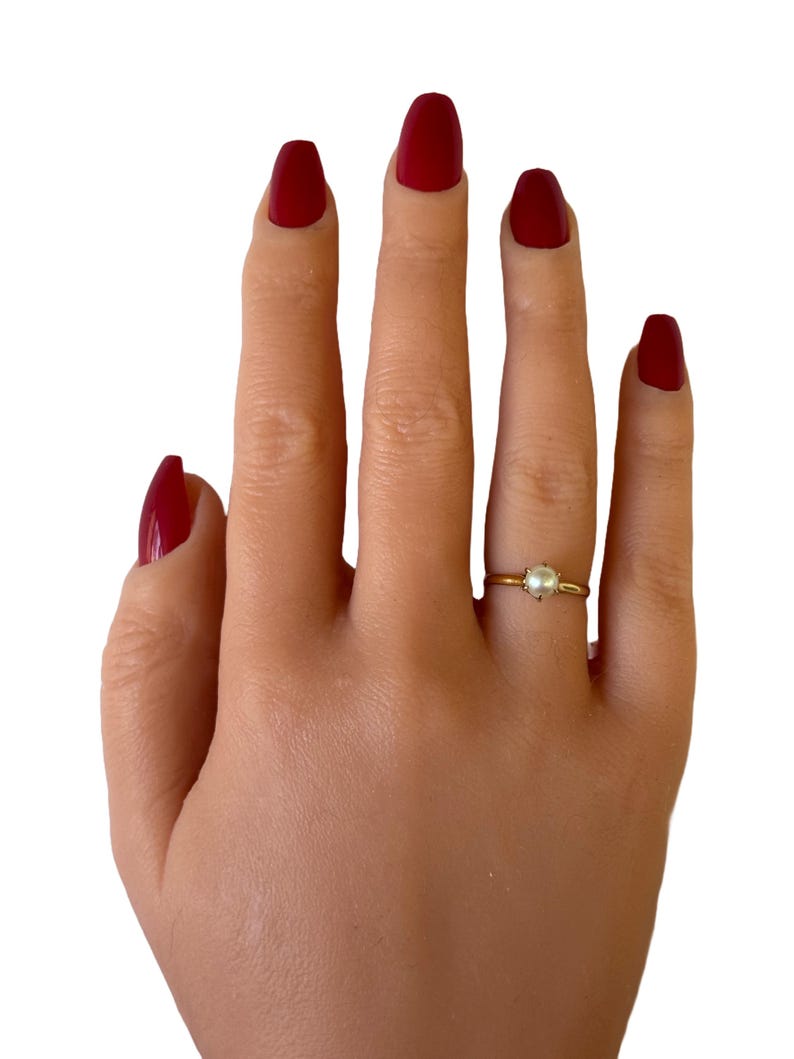May include: A gold ring with a single white pearl on a hand with red nail polish.