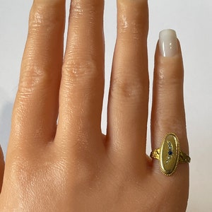 Peut inclure: Une bague en or avec un chaton de forme ovale serti d'une pierre bleue et de petites pierres blanches. La bague a une bande textur&eacute;e et d&eacute;taill&eacute;e.