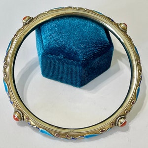 K&ouml;nnte beinhalten: Ein silbernes Armreif-Armband mit blauen und roten Emaildetails. Das Armband ist mit kleinen, runden, roten Steinen verziert.