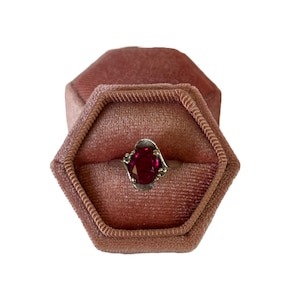 May include: A red gemstone ring in a pink velvet ring box. The ring has a silver band and an oval-shaped red gemstone.