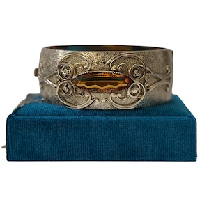 Puede incluir: Brazalete rígido plateado con una piedra ovalada de color ámbar. El brazalete presenta adornos de volutas y se exhibe sobre una caja de terciopelo azul. Una joya.