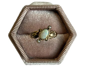 Op de afbeelding: Een gouden ring met een ovale opaal edelsteen en kleine parel accenten, gezet in een decoratieve band. De ring wordt gepresenteerd in een zeshoekige, lichtroze fluwelen sieradendoos. Het ontwerp van de ring heeft sierlijke details.