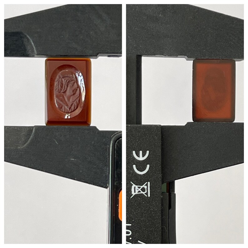 Op de afbeelding: Een close-up van een rode carneol cameo met een gegraveerd profiel van een vrouwengezicht. De cameo wordt gemeten met een schuifmaat, wat de grootte van de steen laat zien.