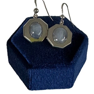 May include: A pair of silver earrings with octagonal frames and white stones. The earrings are displayed on a blue velvet cushion.