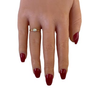 May include: A simple gold ring with a small pearl in the center. The ring is on a finger with red nail polish.