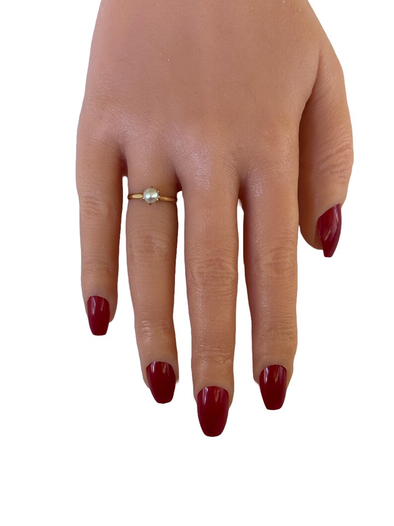 May include: A simple gold ring with a small pearl in the center. The ring is on a finger with red nail polish.