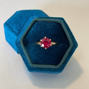 May include: A red gemstone ring in a teal velvet ring box.