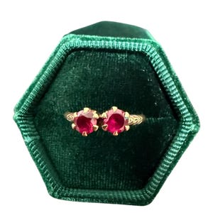 May include: A gold ring with two round, bright red gemstones, set in a decorative filigree design. The ring is displayed in a dark green, hexagonal velvet ring box, creating a striking contrast. The gemstones are the focal point.