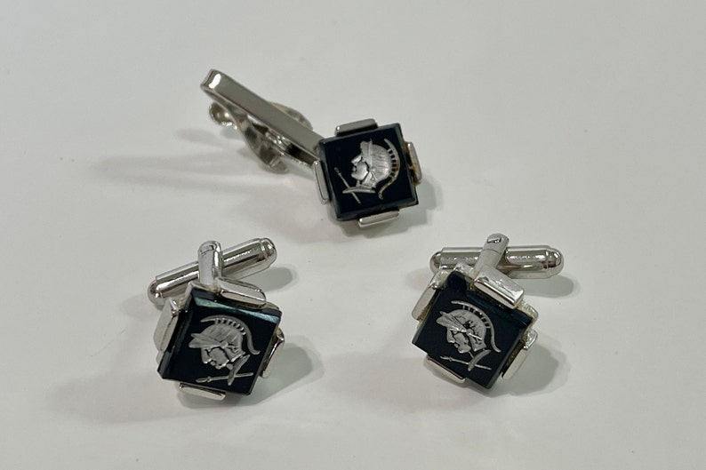 May include: A set of silver cufflinks and a tie clip with black square onyx stones featuring a silver etched design of a Roman soldier's head in profile.