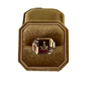 May include: A gold ring with a red stone and a Masonic symbol. The ring is displayed in a brown velvet ring box.