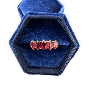 May include: A gold ring featuring five oval-cut red gemstones, arranged in a line. The ring is presented in an open, hexagonal blue velvet jewellery box. The gemstones are a vivid red, and the gold setting offers a warm contrast.