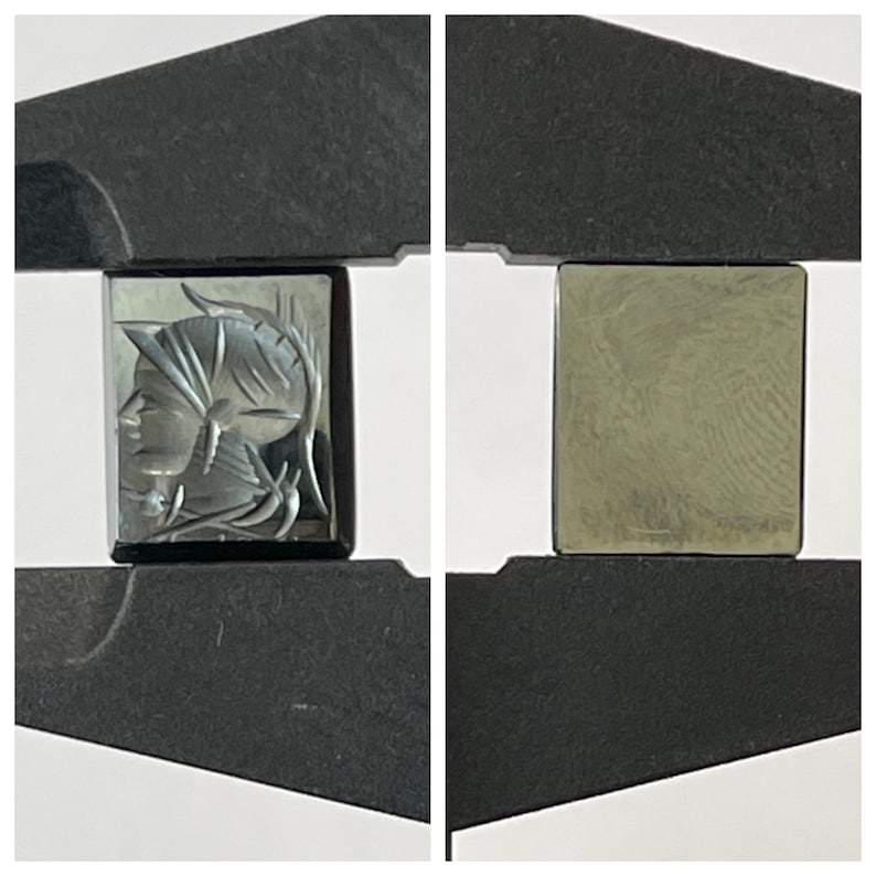 Op de afbeelding: Een zwarte en zilveren gegraveerde edelsteen met een profiel van een menselijk gezicht. De edelsteen wordt vastgehouden in een metalen klem.