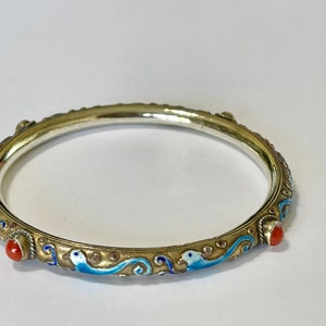 K&ouml;nnte beinhalten: Ein silbernes Armreif-Armband mit blauen und wei&szlig;en Emaildetails und roten Korallenakzenten. Das Armband ist mit einem sich wiederholenden Muster aus stilisierten V&ouml;geln und Wellen verziert.