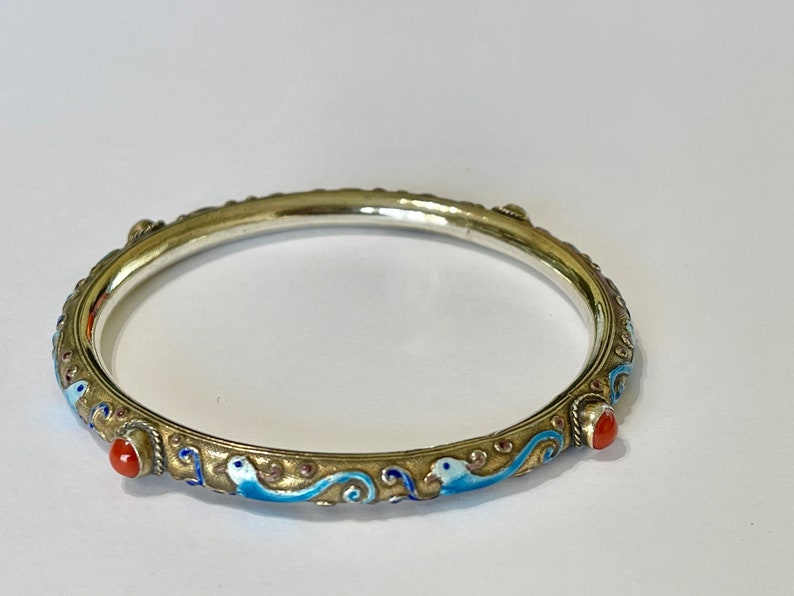 K&ouml;nnte beinhalten: Ein silbernes Armreif-Armband mit blauen und wei&szlig;en Emaildetails und roten Korallenakzenten. Das Armband ist mit einem sich wiederholenden Muster aus stilisierten V&ouml;geln und Wellen verziert.
