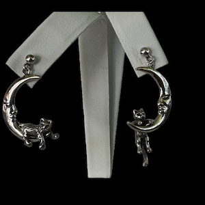 May include: A pair of silver earrings featuring a crescent moon with a cat's face and a cat hanging from the moon.