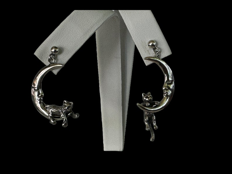 May include: A pair of silver earrings featuring a crescent moon with a cat's face and a cat hanging from the moon.