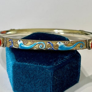 K&ouml;nnte beinhalten: Ein silbernes Armreif-Armband mit blauen und goldenen Emaille-Details und zwei roten Korallensteinen.