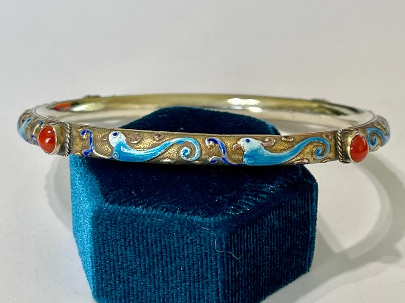 K&ouml;nnte beinhalten: Ein silbernes Armreif-Armband mit blauen und goldenen Emaille-Details und zwei roten Korallensteinen.