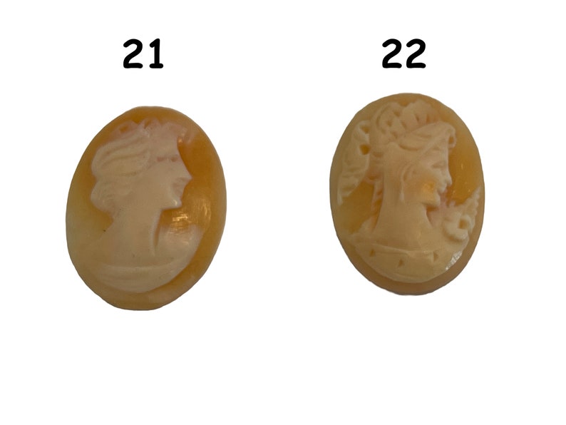 May include: Two oval-shaped cameo pendants with a white cameo image of a woman's profile on a yellow background. The cameo on the left is labeled "21" and the cameo on the right is labeled "22".