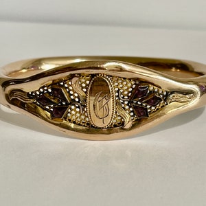 Op de afbeelding: Een goudkleurige armband met een ovale middenstuk met een gegraveerde initiaal. De armband heeft decoratieve filigraan en donkerrode edelsteenaccenten. Het ontwerp bevat bloemen- en bladmotieven.