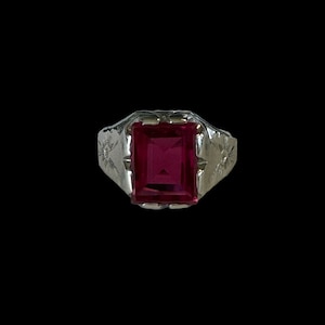Peut inclure: Une bague en argent avec une pierre précieuse rouge carrée sertie au centre. La bague a un motif floral sur la bande.