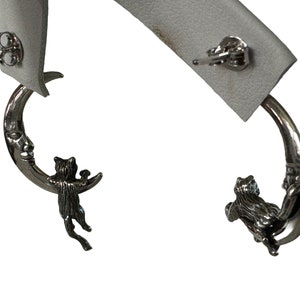 May include: A pair of silver earrings featuring a crescent moon with a cat clinging to it. The cat is holding a small object in its paws.