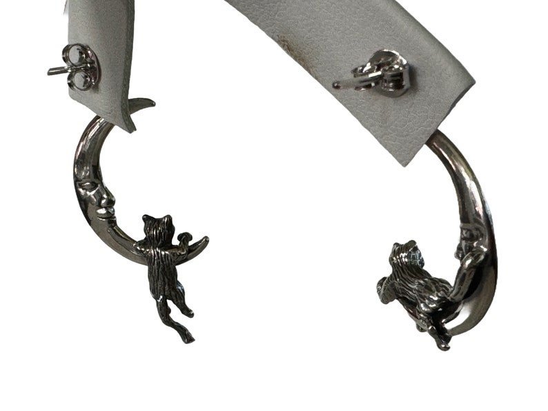 May include: A pair of silver earrings featuring a crescent moon with a cat clinging to it. The cat is holding a small object in its paws.