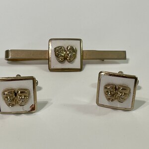 May include: A set of gold-toned cufflinks and a tie bar with white enamel and a design of two gold-toned theatre masks.