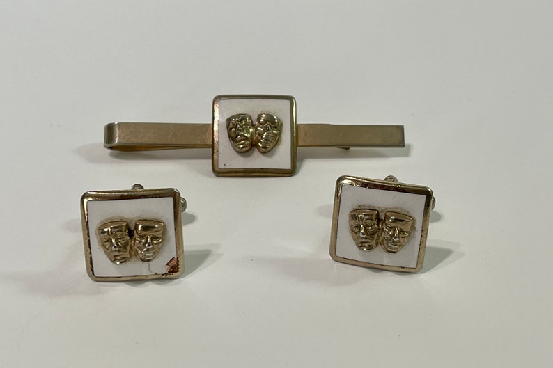 May include: A set of gold-toned cufflinks and a tie bar with white enamel and a design of two gold-toned theatre masks.