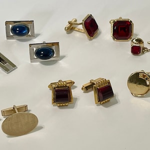 May include: A collection of cufflinks and tie tacks with various designs. Some feature oval blue stones, others have square red stones, and some are plain gold with a circular design.
