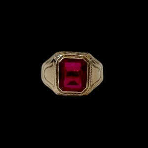 May include: A gold ring featuring a large, rectangular, ruby-red gemstone. The ring has an engraved design around the gemstone and on the band. The background is black, accentuating the ring's details and the gemstone's colour.