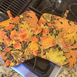 May include: Two quilted pot holders with a fall leaf design in shades of orange, yellow, and green on a black background.