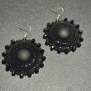 May include: A pair of round, black beaded earrings featuring a central matte black cabochon surrounded by concentric circles of small black seed beads and faceted black crystals, finished with silver-toned metal earring hooks for hanging.