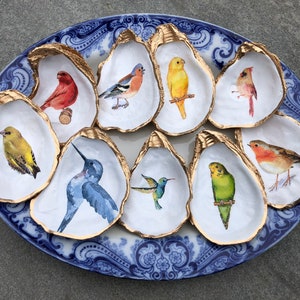 May include: Nine oyster shells painted with gold trim and different bird illustrations. The birds are painted in shades of red, yellow, blue, green, and brown.