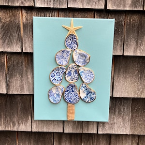 May include: A handmade Christmas tree art piece on a light blue canvas. The tree is crafted from blue and white patterned ceramic pieces with gold trim, topped with a starfish. The trunk is made of cork.